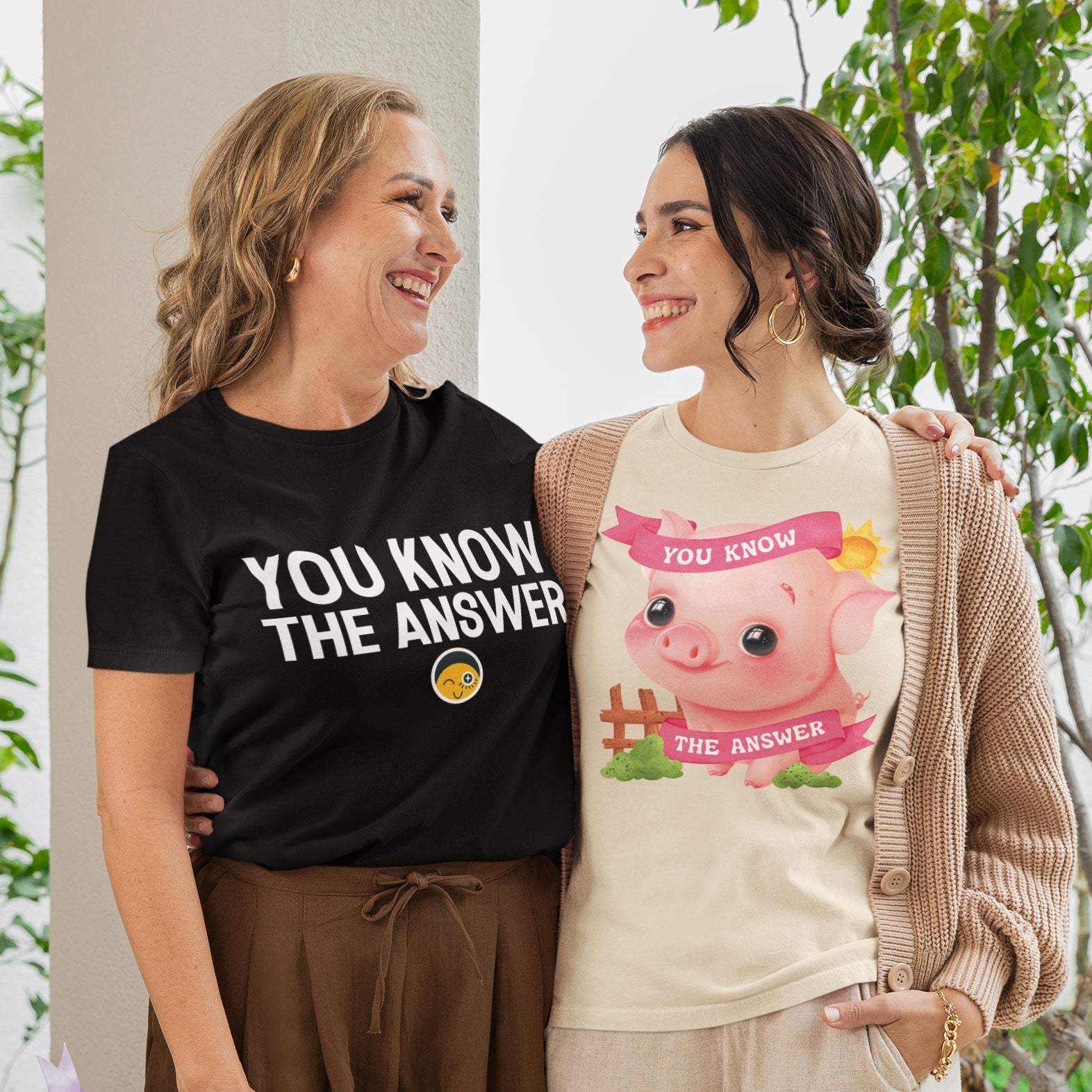 Two women standing together, smiling and posing for a photo. One woman is wearing a black t-shirt with the text "YOU KNOW THE ANSWER" and the other is wearing a t-shirt with a cartoon pig and the text "YOU KNOW THE ANSWER".
