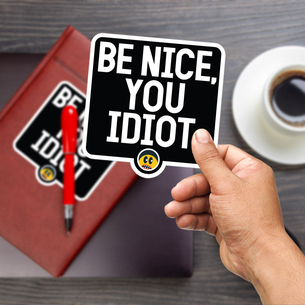 A person's hand is holding a sticker with the text "BE NICE, YOU IDIOT" on it, with a red pen and a laptop visible in the background.