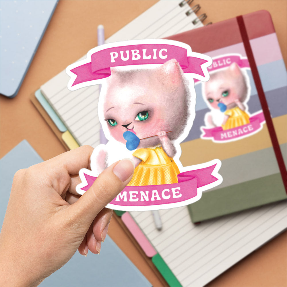 A hand is holding a sticker featuring a cartoon cat with a pink ribbon that says "PUBLIC MENACE".