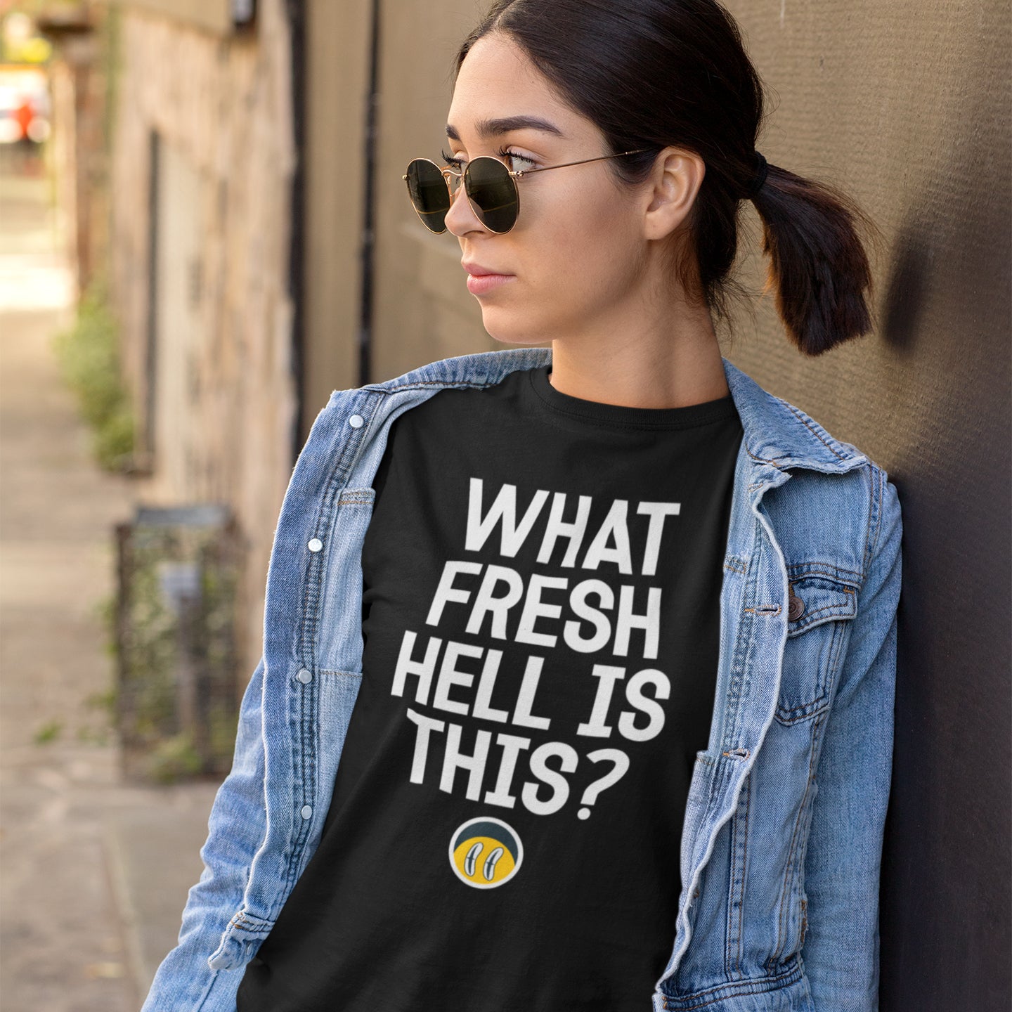A woman wearing a denim jacket and sunglasses stands against a brick wall, wearing a black t-shirt with the text "WHAT FRESH HELL IS THIS?"