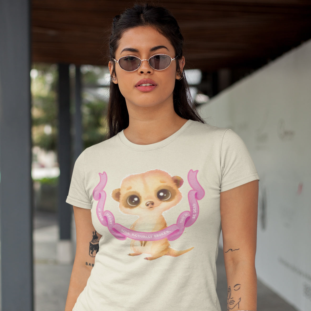 A woman wearing a white t-shirt with a cartoon meerkat graphic and sunglasses stands in front of a building.