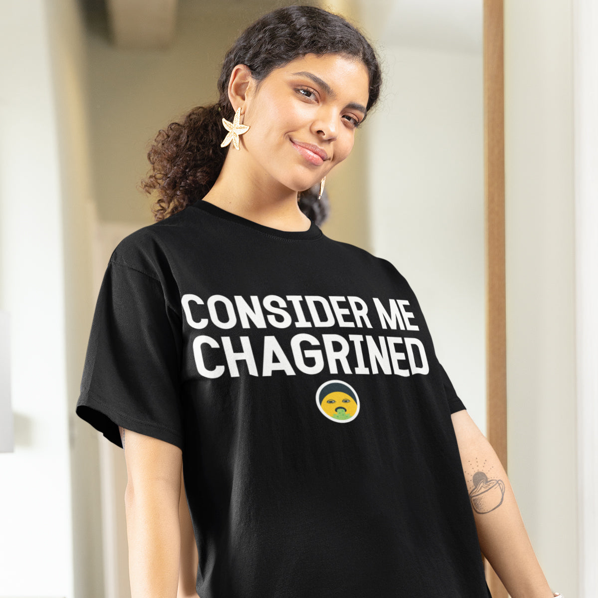 A woman wearing a black t-shirt with the text "CONSIDER ME CHAGRINED" printed on it, smiling at the camera.