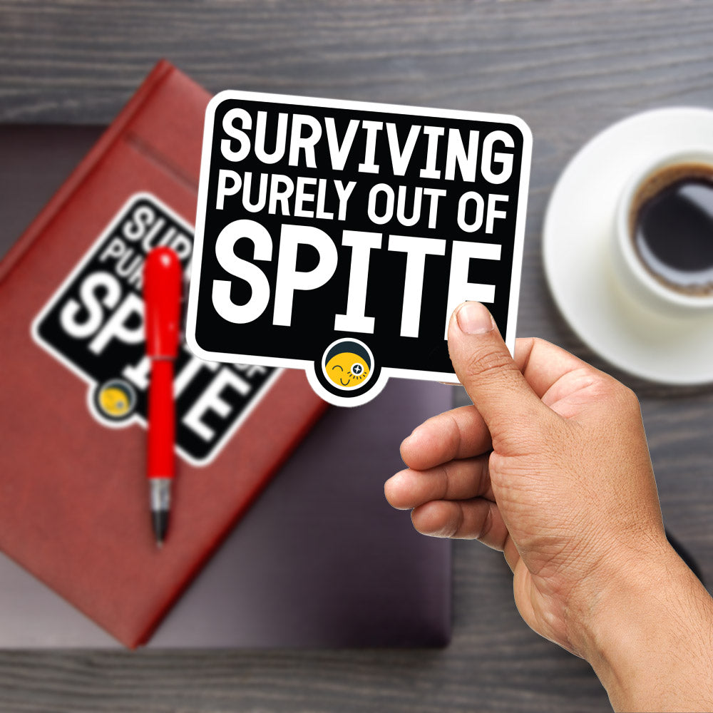 A person's hand is holding a sticker with the text "SURVIVING PURELY OUT OF SPITE" on a wooden surface. There is also a red pen and a cup of coffee visible in the background.