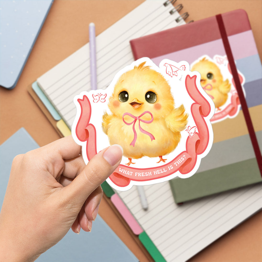 A hand is holding a sticker featuring a cute yellow chick with a bow, surrounded by a ribbon and the text "What fresh hell is this?".