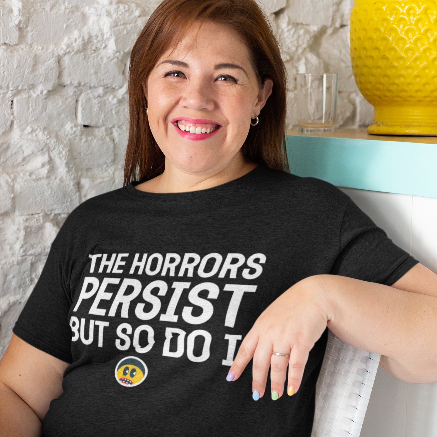 A woman wearing a black t-shirt with white text and a yellow smiley face sticker is smiling and leaning against a white wall.