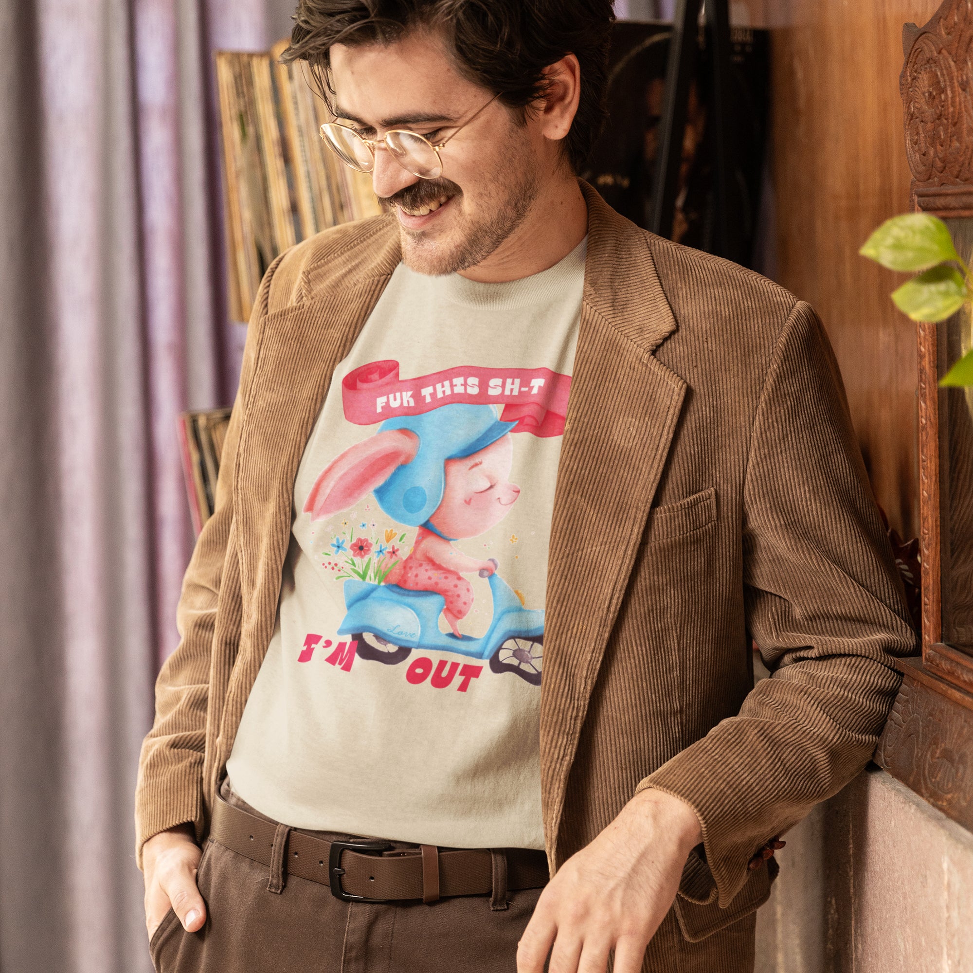 A man wearing a brown jacket and glasses is leaning against a wall, smiling and looking down. He is wearing a t-shirt with a graphic design featuring a cartoon character and the text "I'm out".