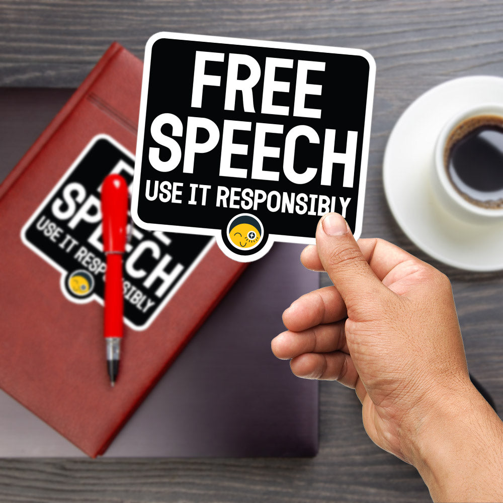A person's hand is holding a sign that says "FREE SPEECH USE IT RESPONSIBLY" with a smiley face sticker. The sign is placed on a wooden surface, and there is a red pen and a cup of coffee nearby.