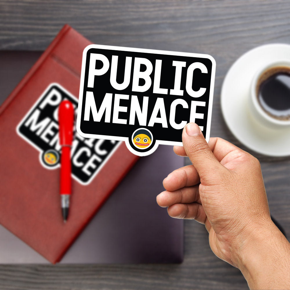 A hand holding a sticker with the text "PUBLIC MENACE" on a wooden surface, with a red pen and a book with a sticker on it in the background.