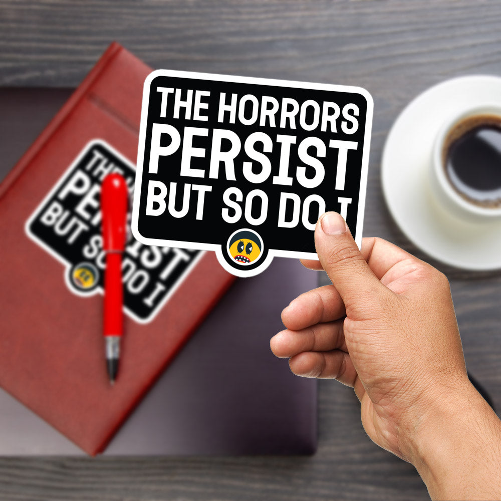A person's hand is holding a sticker with the text "The Horrors Persist but So Do!" against a wooden background. There is also a red pen and a cup of coffee visible in the image.