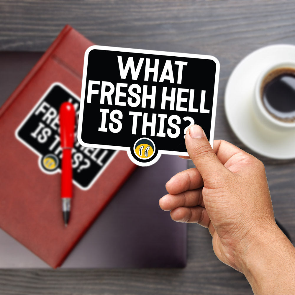 A hand holding a sticker with the text "WHAT FRESH HELL IS THIS?" in front of a laptop and a cup of coffee on a wooden surface.
