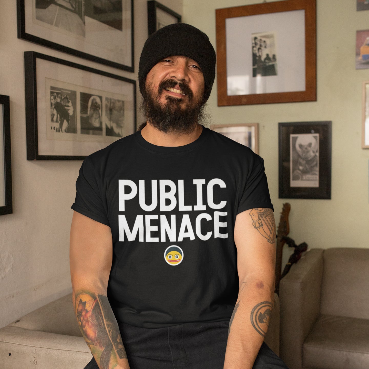 A man with a beard wearing a black t-shirt with the text "PUBLIC MENACE" is sitting in a room with framed pictures on the walls.
