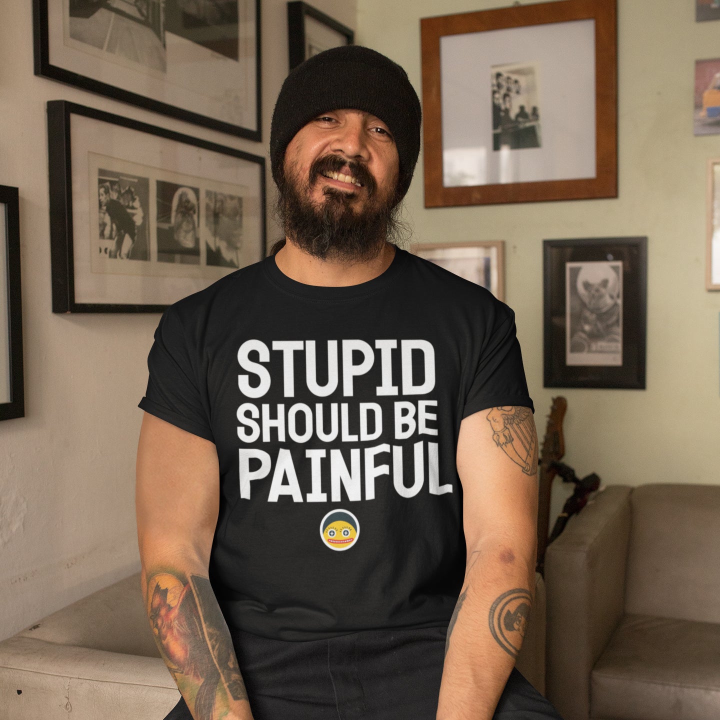 A man with a beard wearing a black t-shirt with the text "STUPID SHOULD BE PAINFUL" is sitting in a room with framed pictures on the wall.