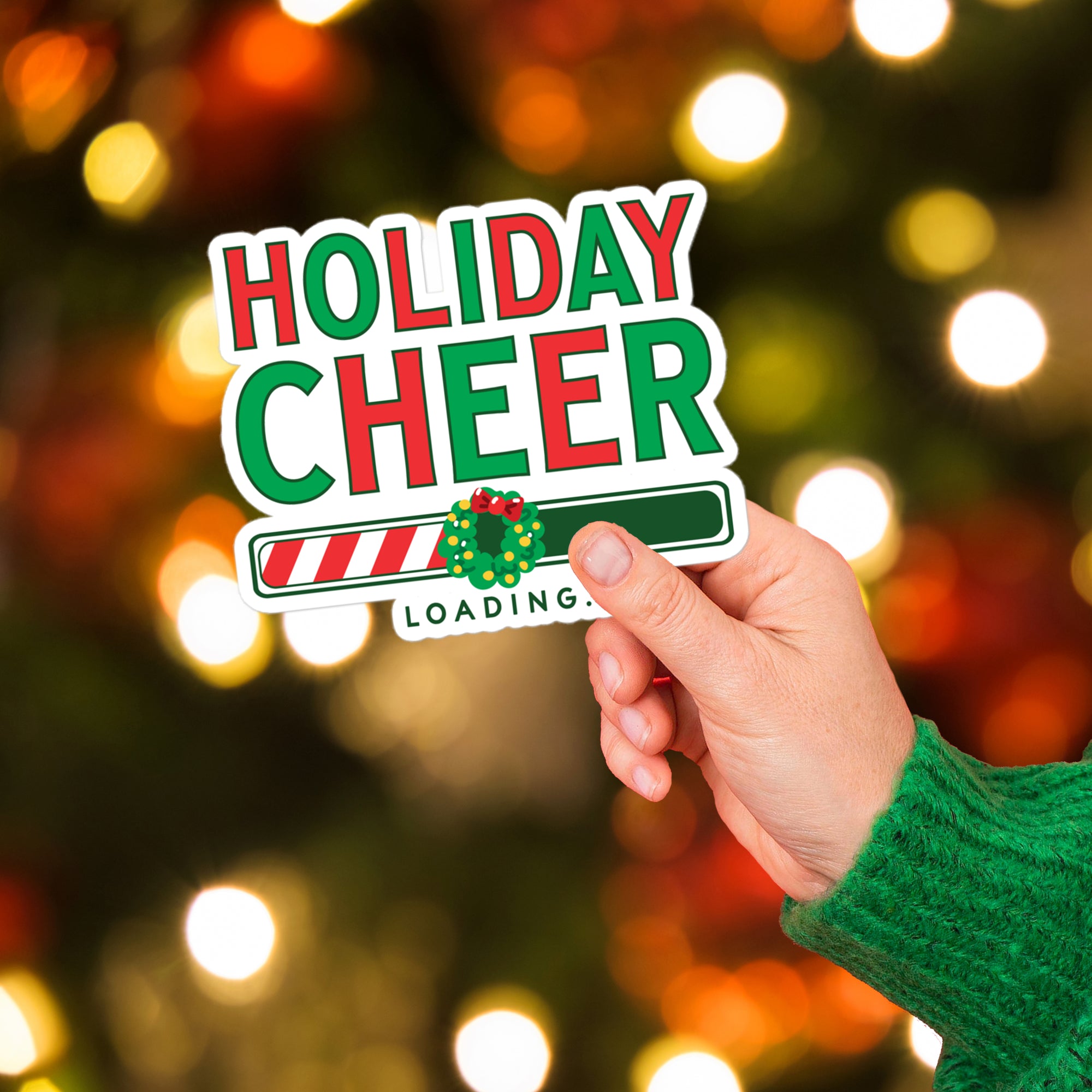 a hand holding a holiday cheer sticker in front of a christmas tree