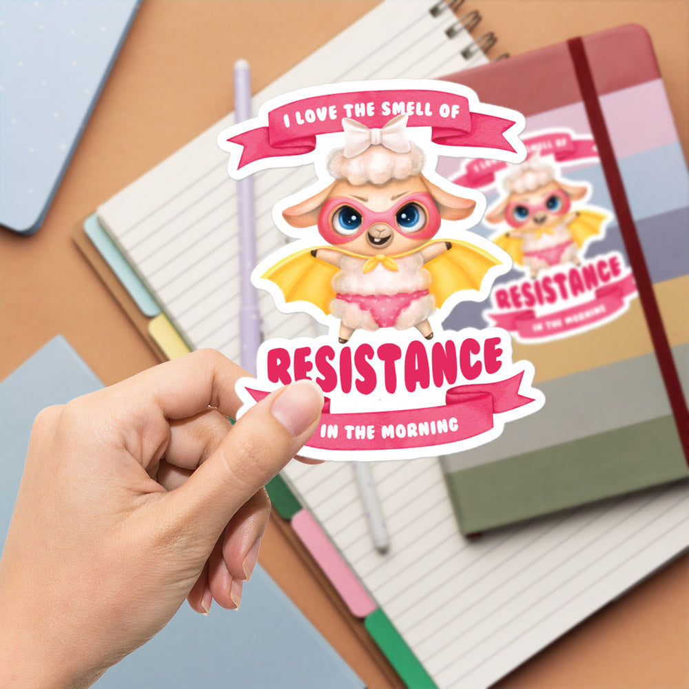 A hand is holding a sticker featuring a cartoon character with wings and a bow, along with the text "I LOVE THE SMELL OF RESISTANCE IN THE MORNING".