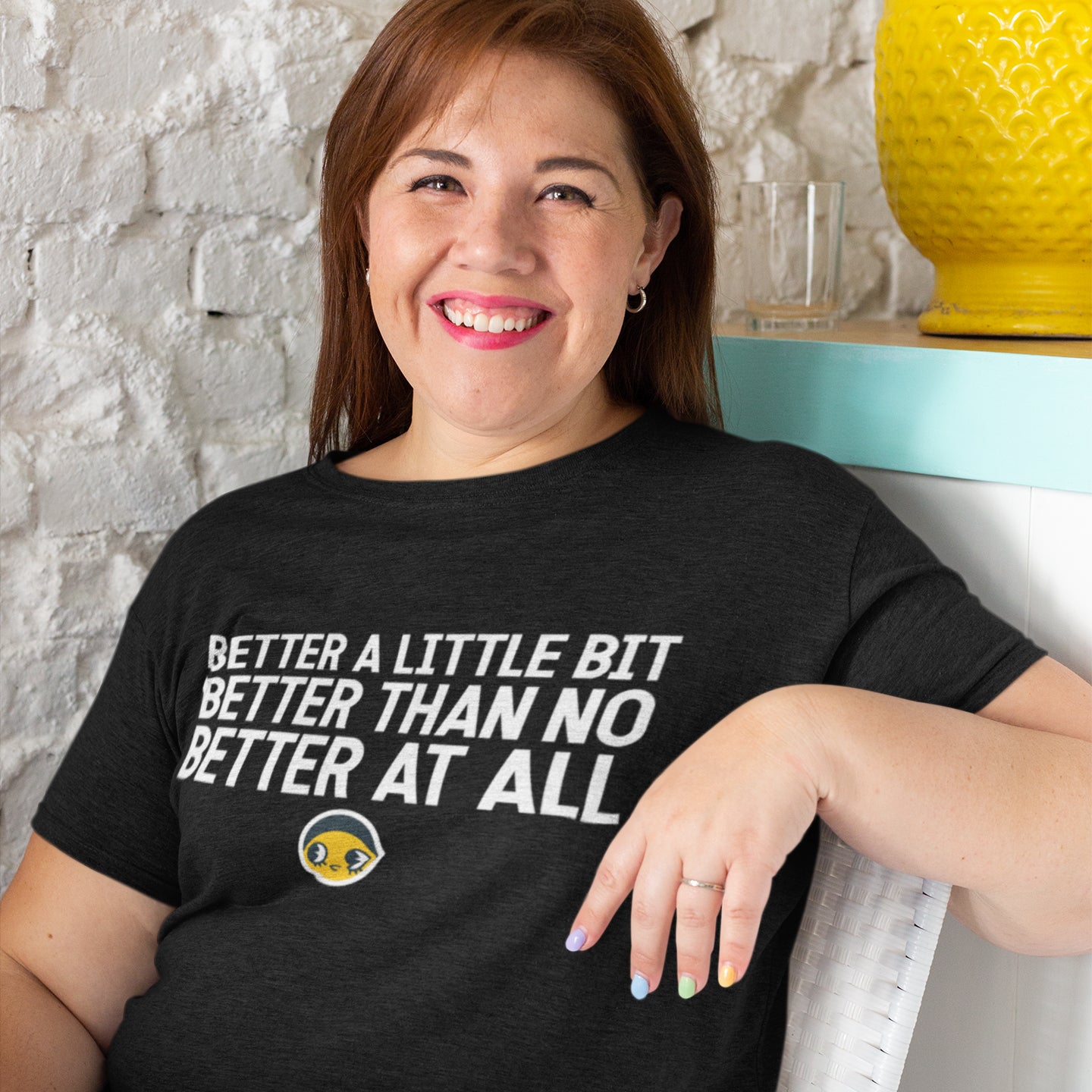 A woman wearing a black t-shirt with the text "Better a little bit better than no better at all" is smiling and leaning against a wall.
