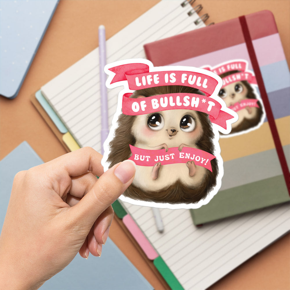 A hand is holding a sticker with a cartoon hedgehog and the text "Life is full of bullsh*t but just enjoy!"