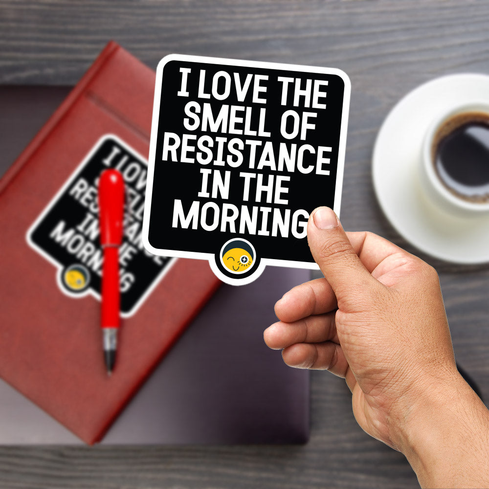A person's hand holding a sticker with the text "I LOVE THE SMELL OF RESISTANCE IN THE MORNING" and a small smiley face sticker.