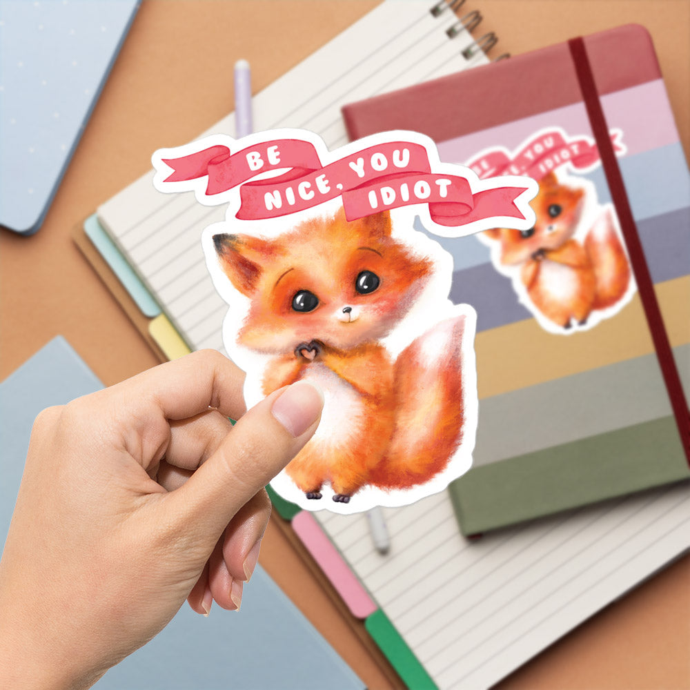A hand is holding a sticker with a cartoon image of a cat and the text "Be nice, you idiot" written on it.