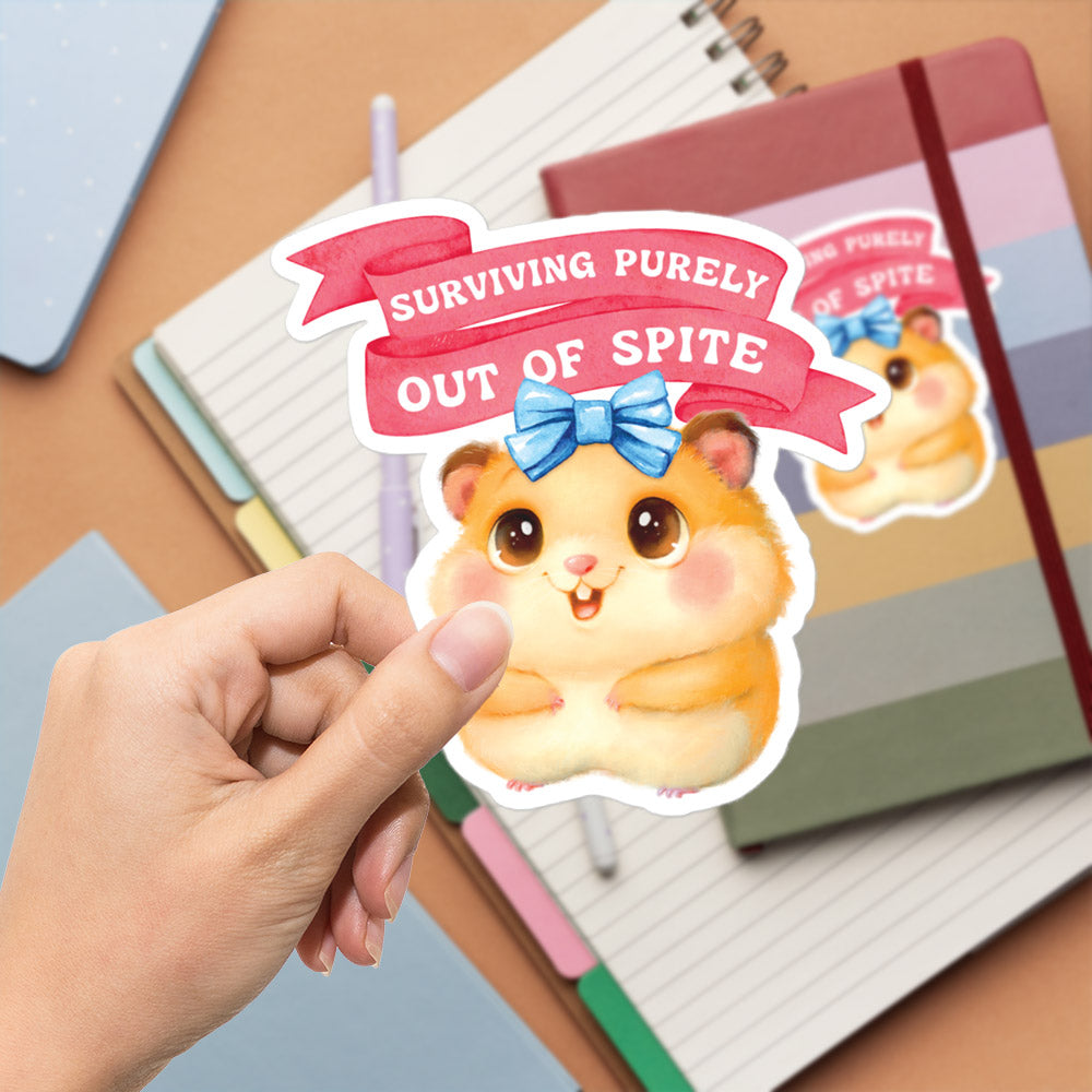 A hand is holding a sticker featuring a cartoon hamster with a bow on its head, surrounded by office supplies such as notebooks and pens.