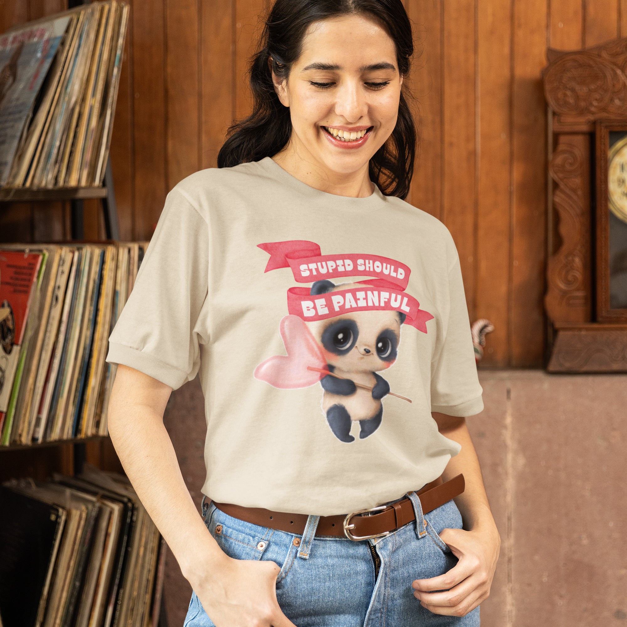 A woman wearing a t-shirt with a cartoon panda and the text "Stupids should be Painful" is smiling and posing in front of a bookshelf.
