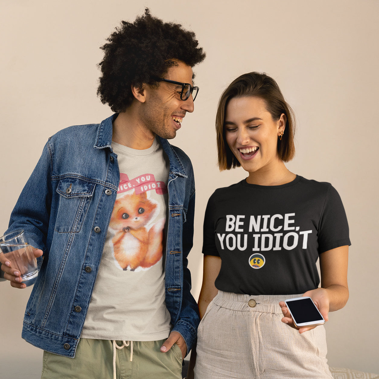 A man and a woman are standing next to each other, smiling and holding drinks. The man is wearing a denim jacket and a t-shirt with a cartoon image, while the woman is wearing a black t-shirt with the text "Be nice, you idiot" printed on it.
