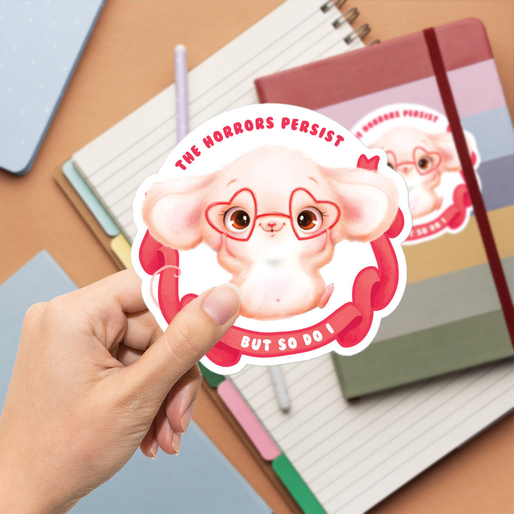 A hand holding a sticker with a cartoon image of a pink mouse wearing glasses and a ribbon, with the text "The Horrors Persist BUT SO DO I" printed on it.