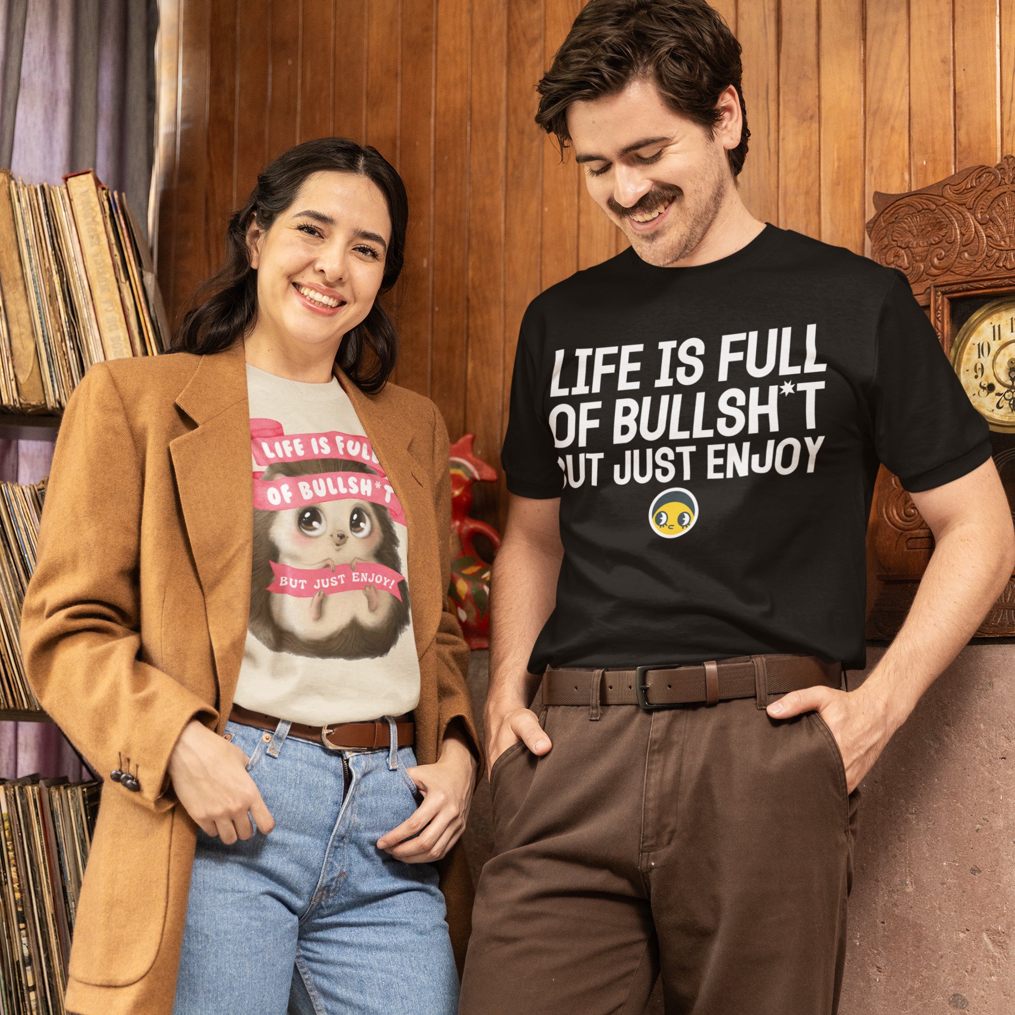 A man and a woman are standing together, smiling and posing for a photo. The man is wearing a black t-shirt with a humorous text, while the woman is wearing a pink t-shirt with a cartoon character. They are standing in front of a bookshelf, which has various books on it.