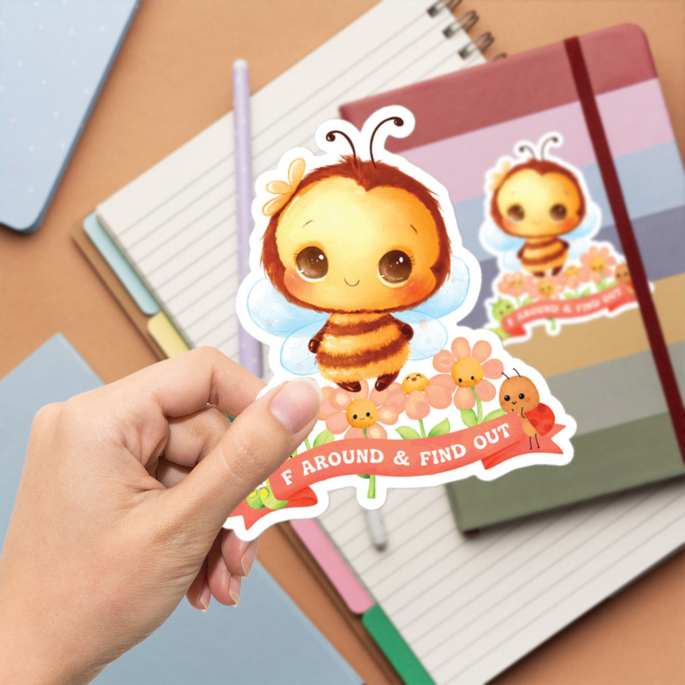 A hand is holding a sticker featuring a cartoon bee surrounded by flowers and the text "F AROUND & FIND OUT".