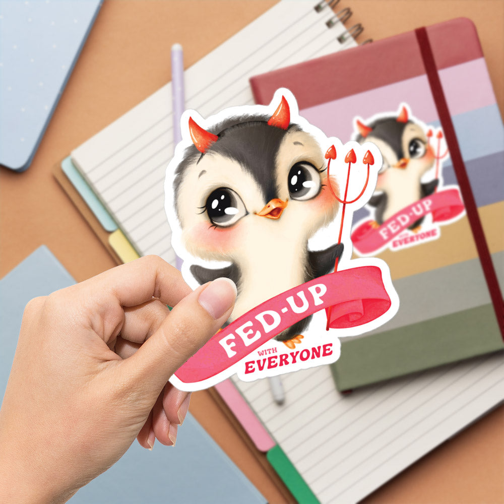 A hand is holding a sticker featuring a cartoon penguin with horns and a banner that says "fed-up with everyone".