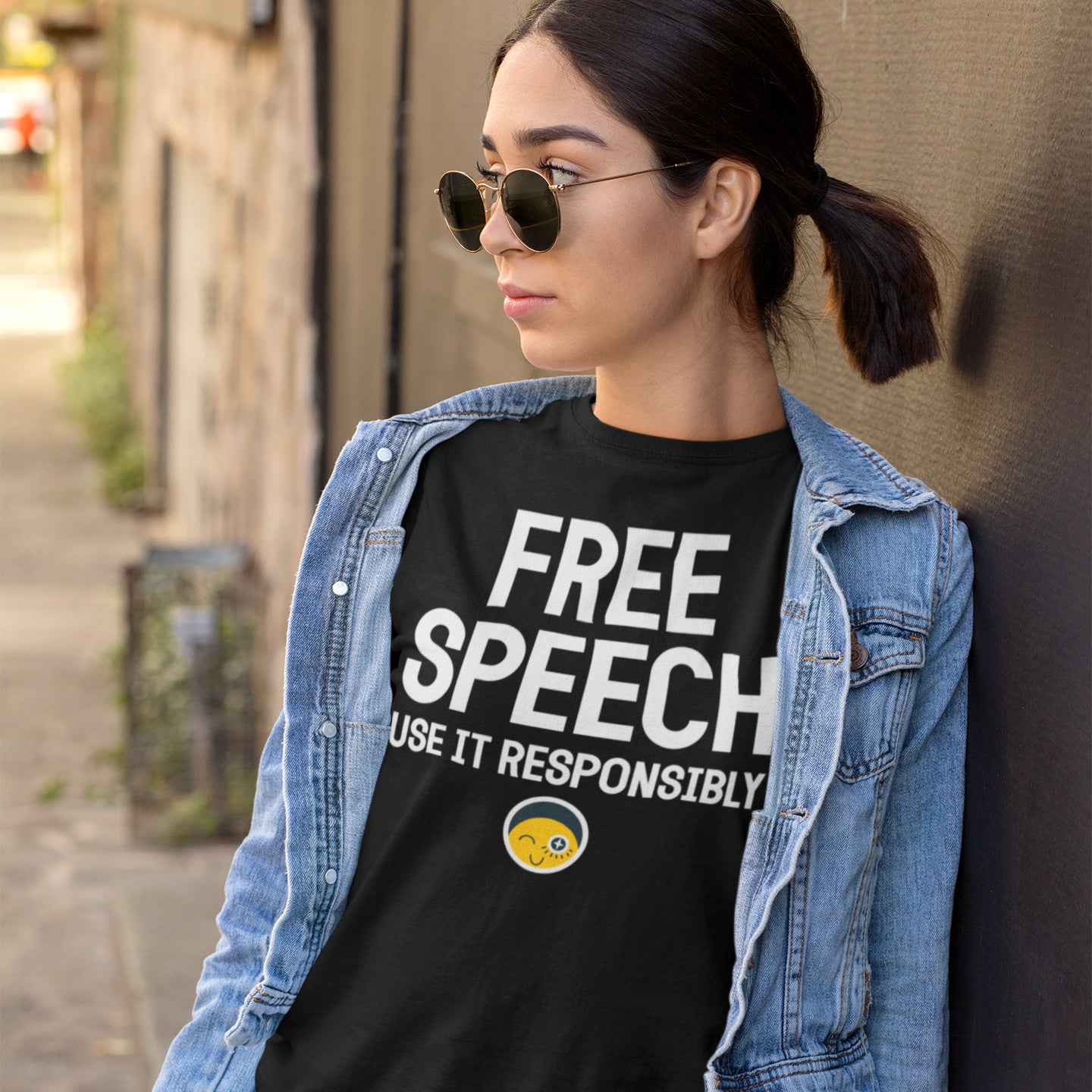 A woman wearing a denim jacket and sunglasses stands against a wall, wearing a black t-shirt with the text "FREE SPEECH USE IT RESPONSIBLY" printed on it.