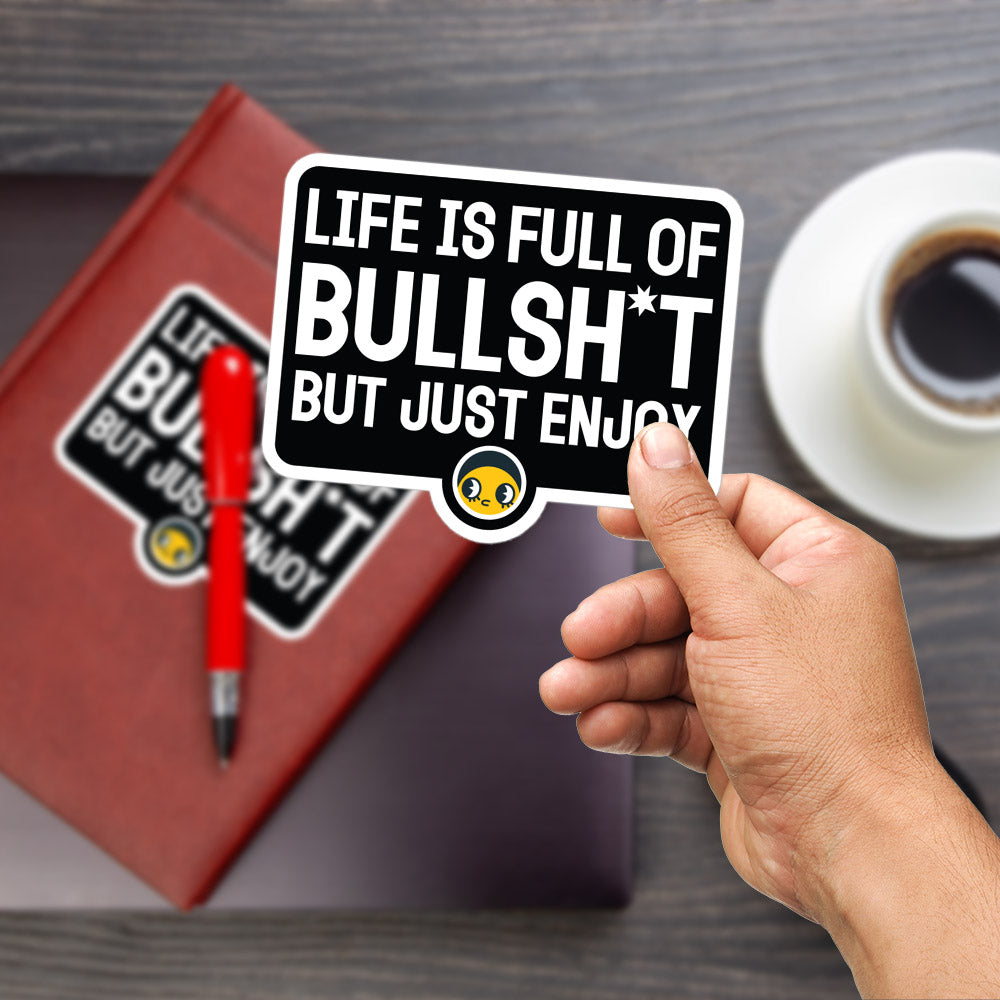A person's hand is holding a sticker with the text "LIFE IS FULL OF BULLSHIT * BUT JUST ENJOY" on it. The hand is also holding a red pen and a laptop, suggesting a workspace or study environment.