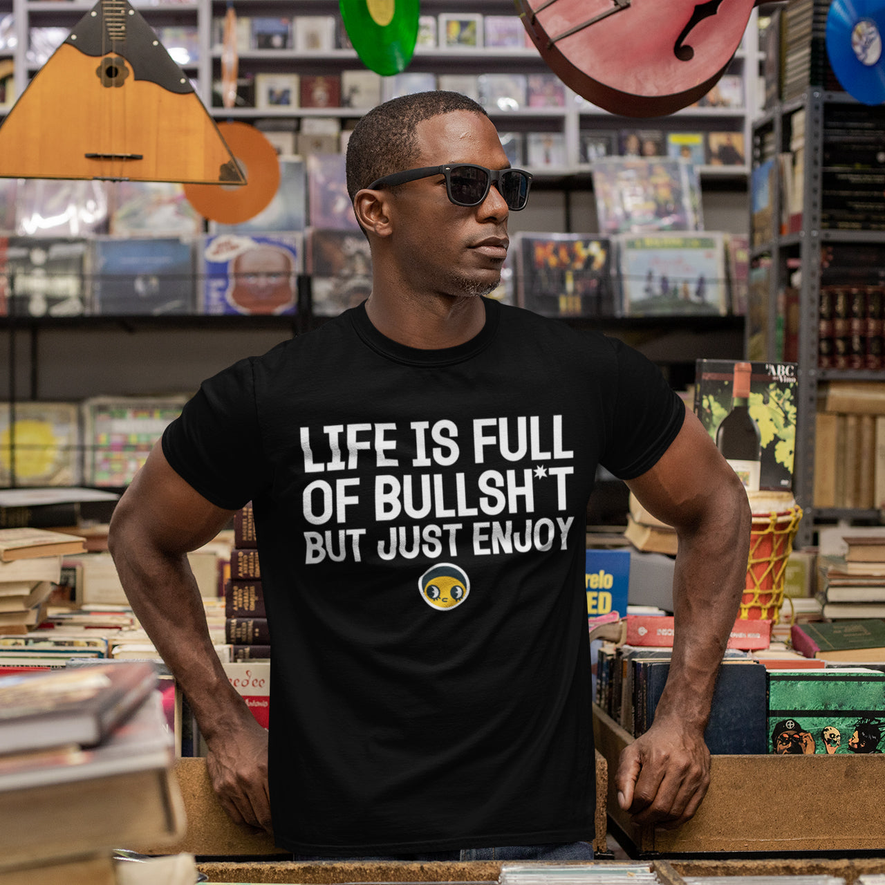 A man wearing a black t-shirt with white text stands in a bookstore surrounded by bookshelves.