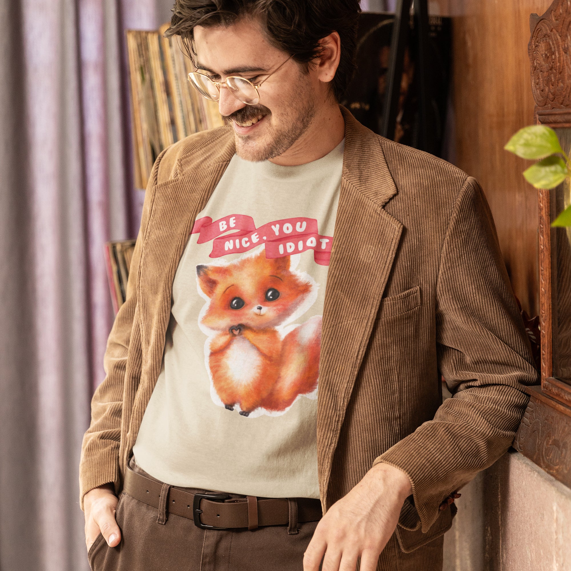 A man wearing a brown jacket and a t-shirt with a cartoon image of a fox is leaning against a wall.