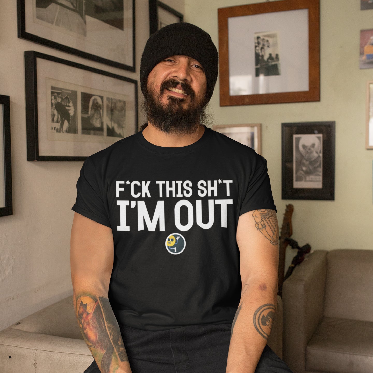 A man with a beard wearing a black beanie and a black t-shirt with white text is sitting in a room with framed pictures on the wall.