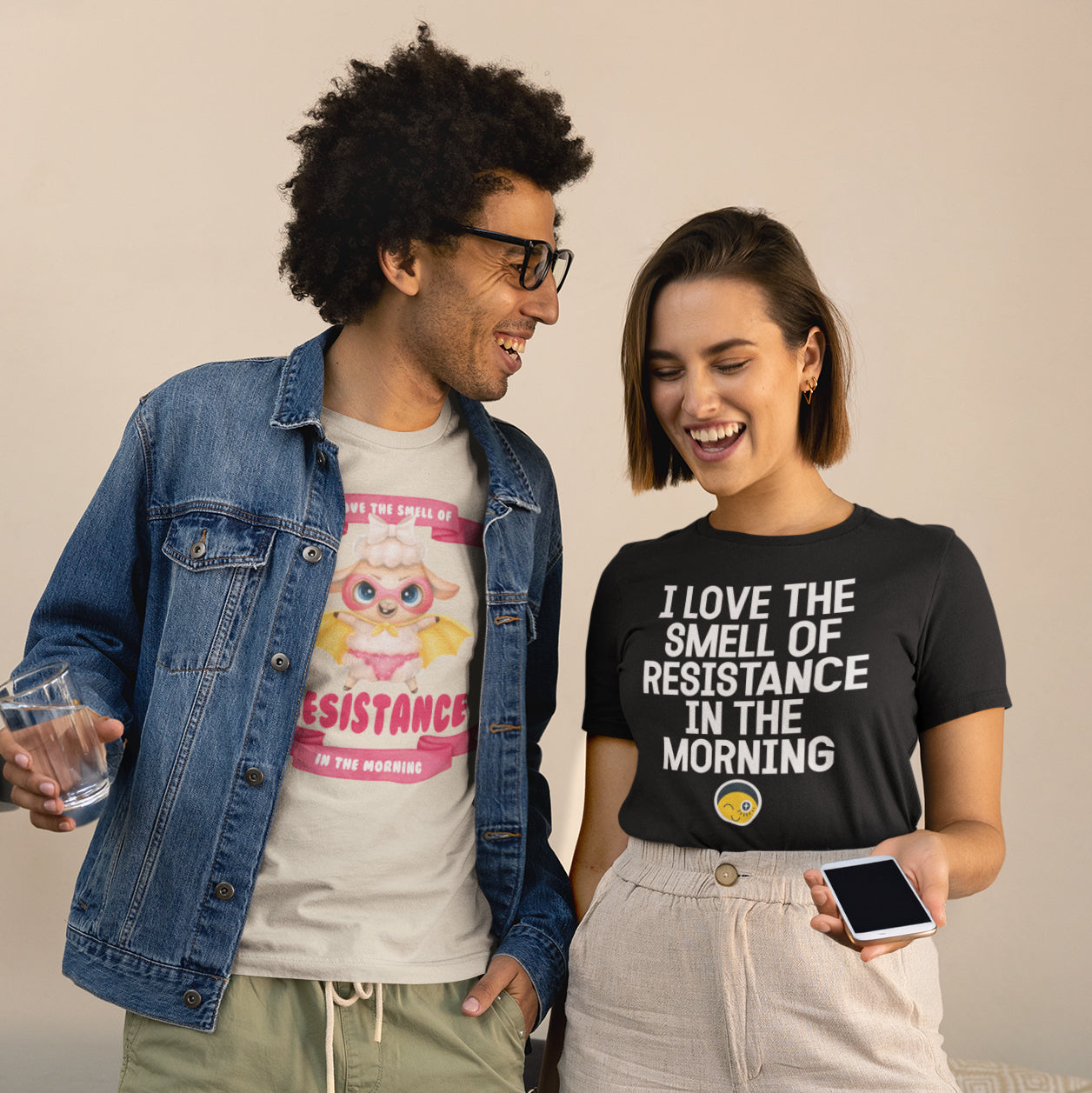 A man and a woman are standing next to each other, smiling and holding a glass and a phone, respectively. They are both wearing casual clothing, including a denim jacket and a black t-shirt with text.