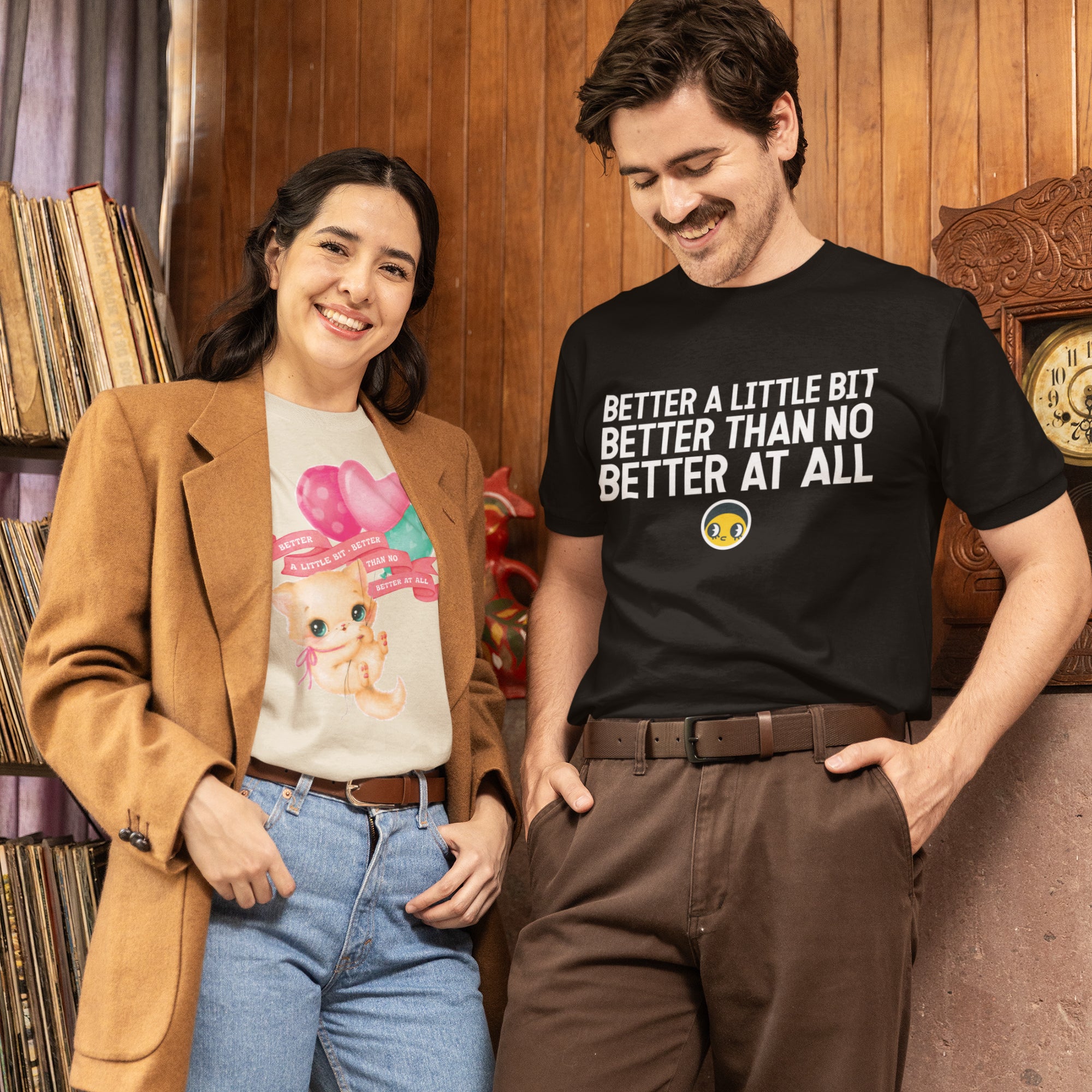 A man and a woman are standing together, smiling and posing for a photo. The man is wearing a black t-shirt with text on it, and the woman is wearing a white t-shirt with a graphic design. They are standing in front of a bookshelf, which has various books on it.