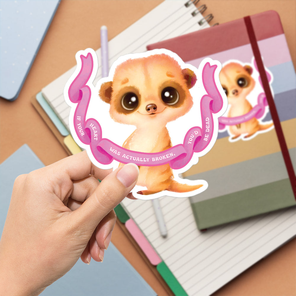 A hand is holding a sticker featuring a meerkat with a pink ribbon around its neck.