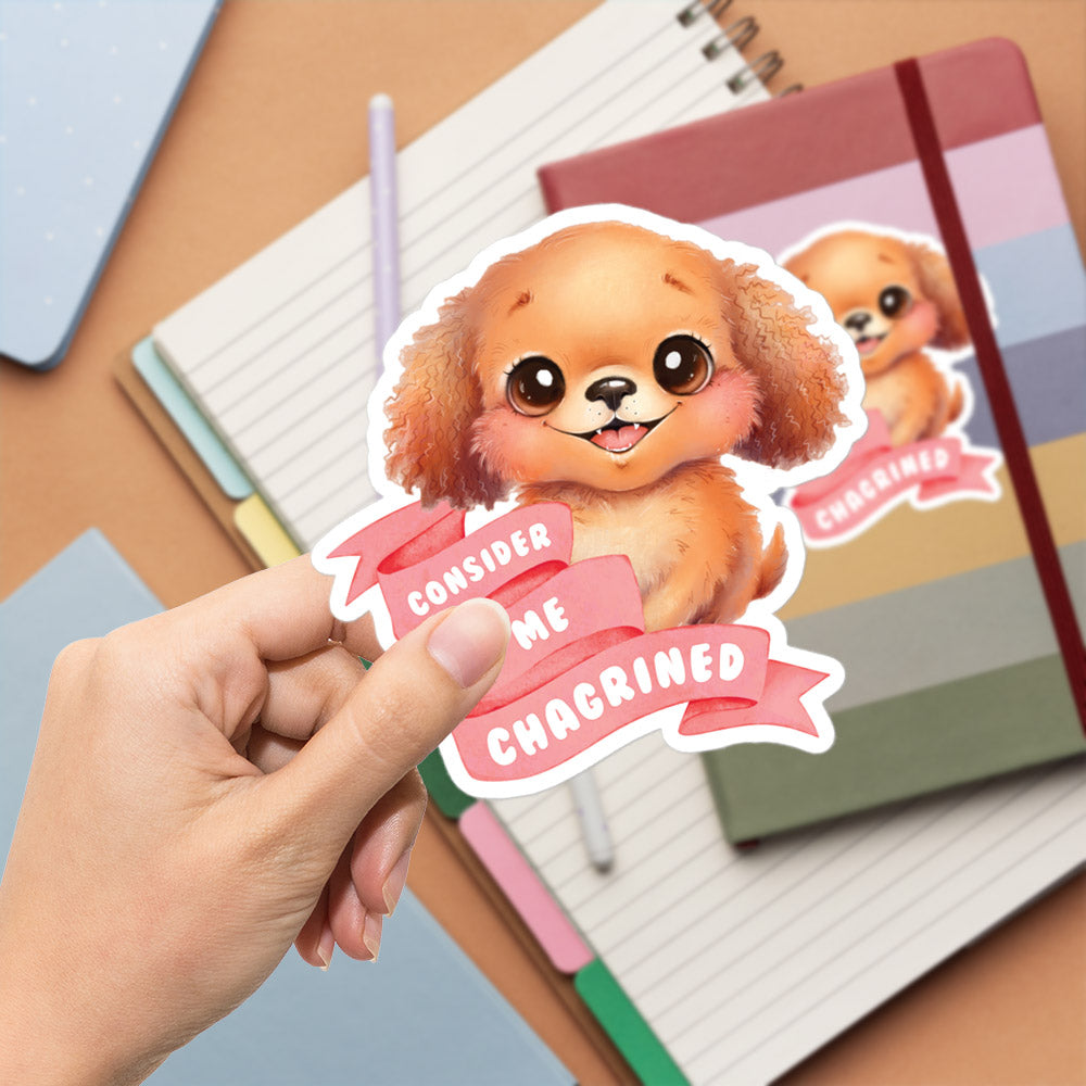 A hand is holding a sticker featuring a cartoon dog with a smiling expression, and the sticker has the text "Considering me Chagrined" written on it.