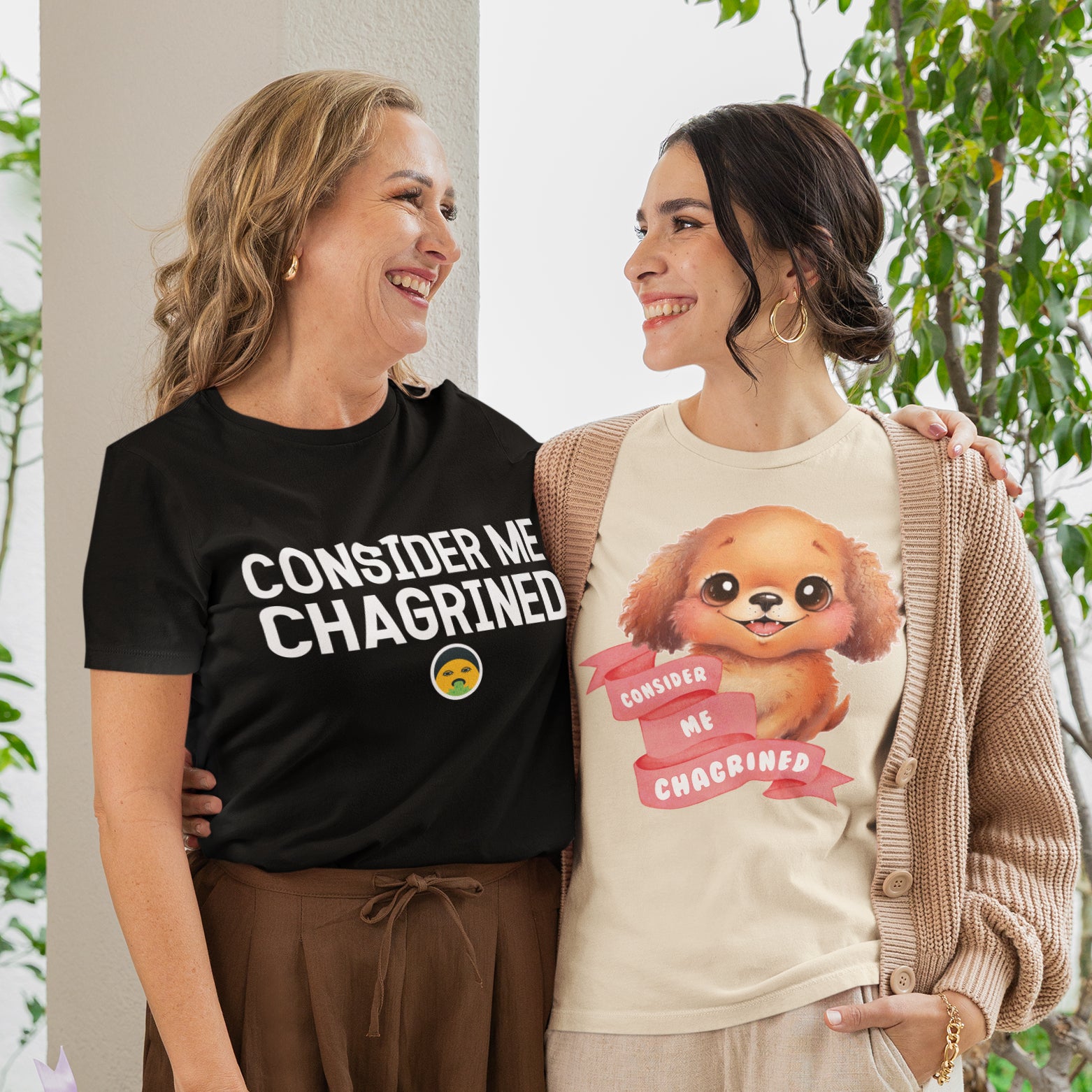 Two women standing together, smiling and posing for a photo. One woman is wearing a black t-shirt with a humorous text, while the other is wearing a t-shirt with a cartoon dog and a pink ribbon with text.