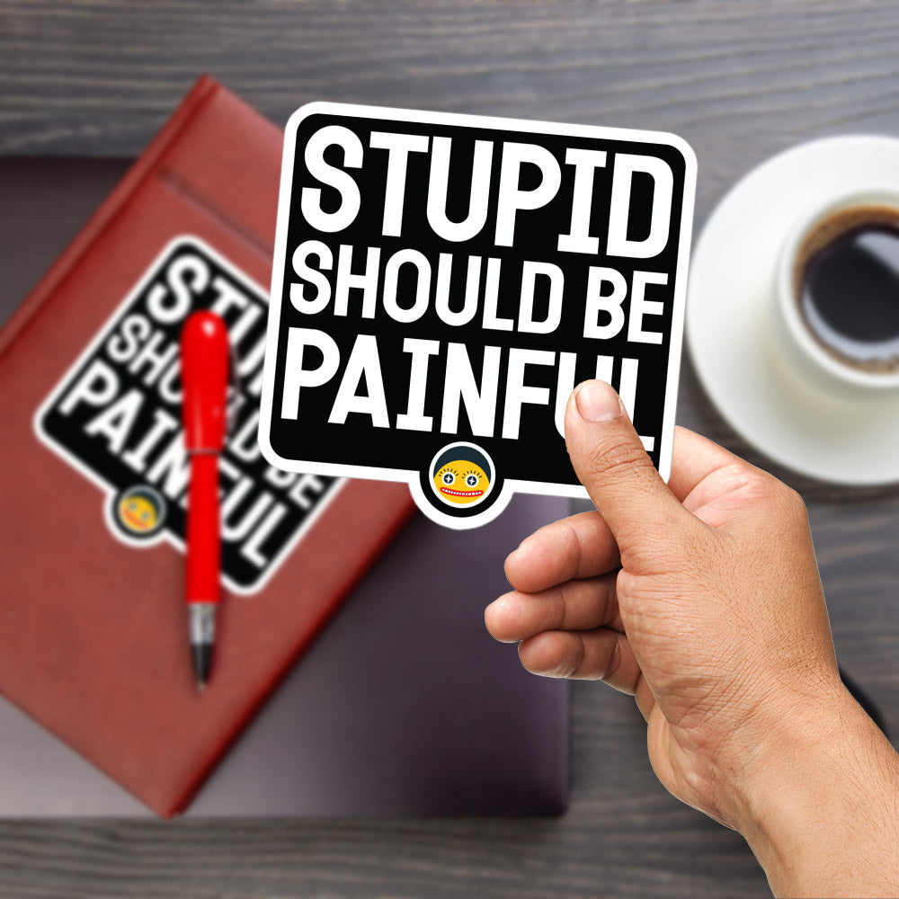 A hand holding a sticker with the text "STUPID SHOULD BE PAINFUL" in bold letters, with a red pen and a book with a sticker on it in the background.