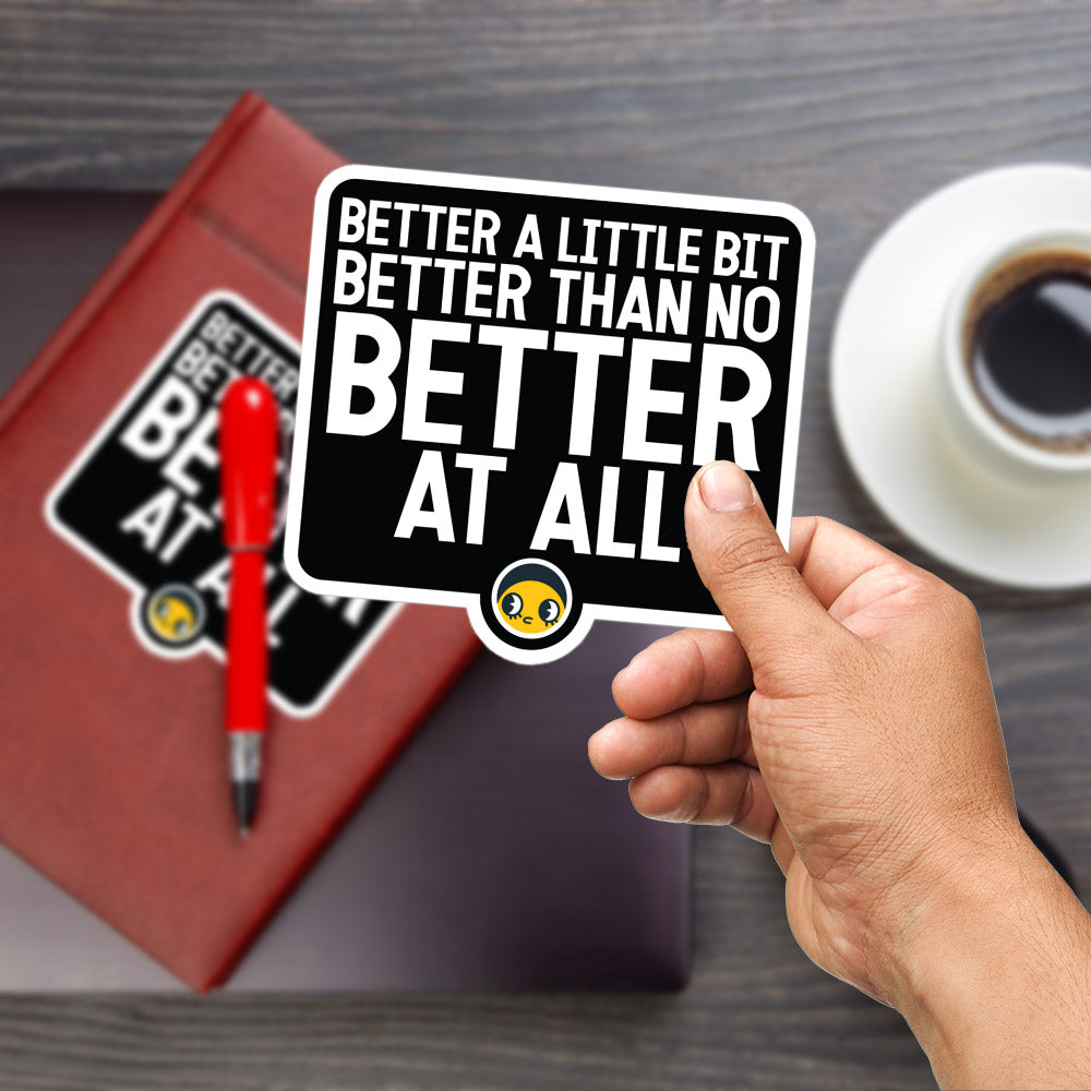 A person's hand holding a sticker with the text "Better a little bit better than no better at all" against a wooden background.