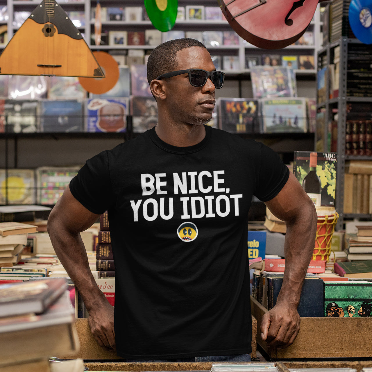 A man wearing a black t-shirt with the text "Be nice, you idiot" stands in a bookstore surrounded by bookshelves.
