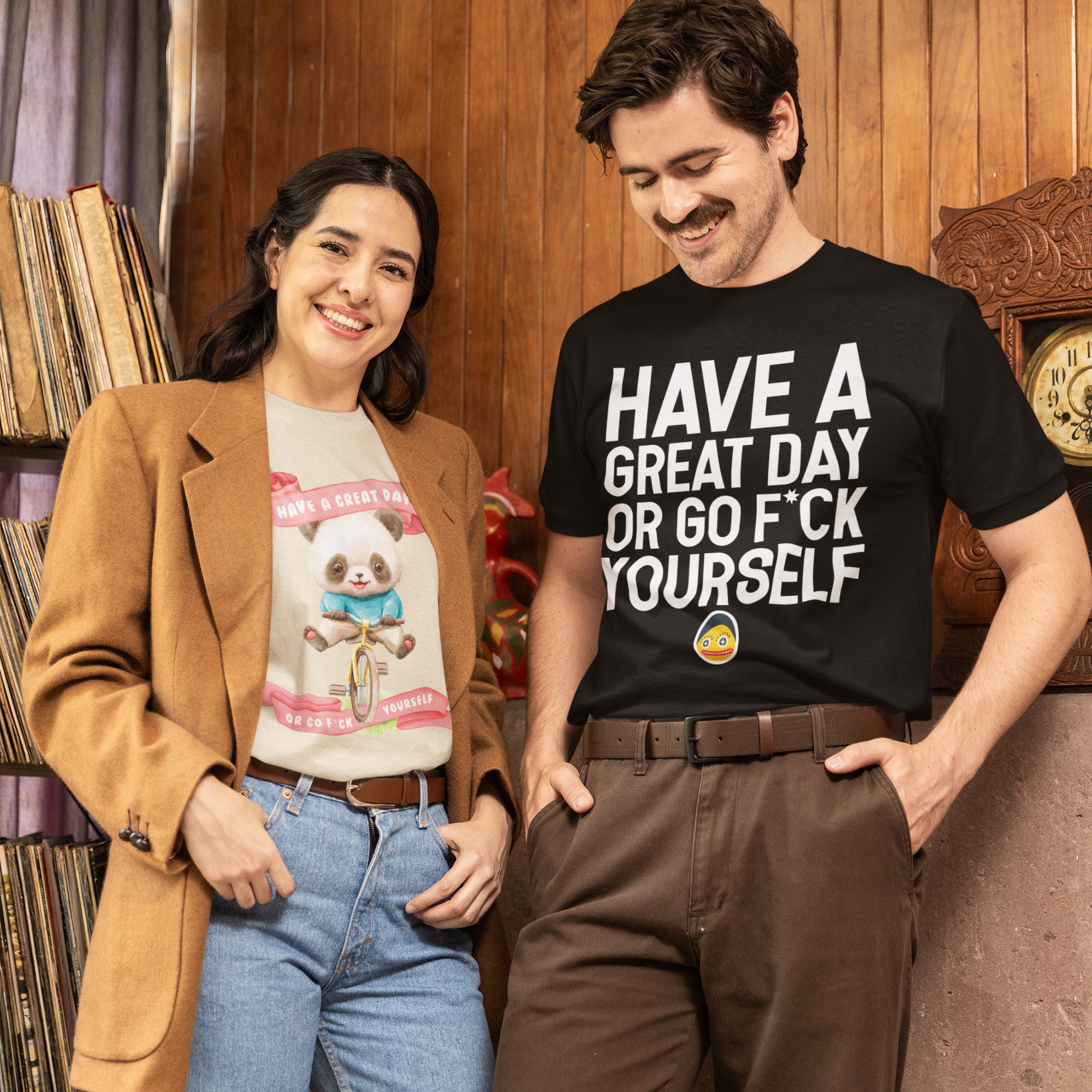 A man and a woman are standing together, smiling and posing for a photo. The man is wearing a black t-shirt with the text "HAVE A GREAT DAY OR GO F*CK YOURSELF" printed on it. The woman is wearing a t-shirt with a cartoon panda and the text "HAVE A GREAT DAY OR GO F*CK YOURSELF" printed on it.