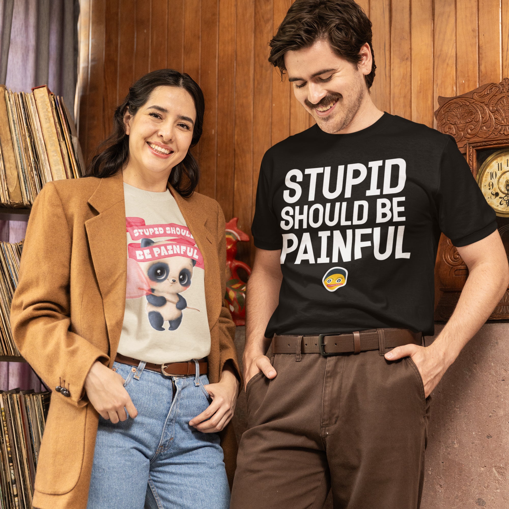A man and a woman are standing together, smiling and posing for a photo. The man is wearing a black t-shirt with the text "STUPID SHOULD BE PAINFUL" printed on it, while the woman is wearing a t-shirt with a cartoon character and the text "STUPID SHOULD BE PAINFUL".