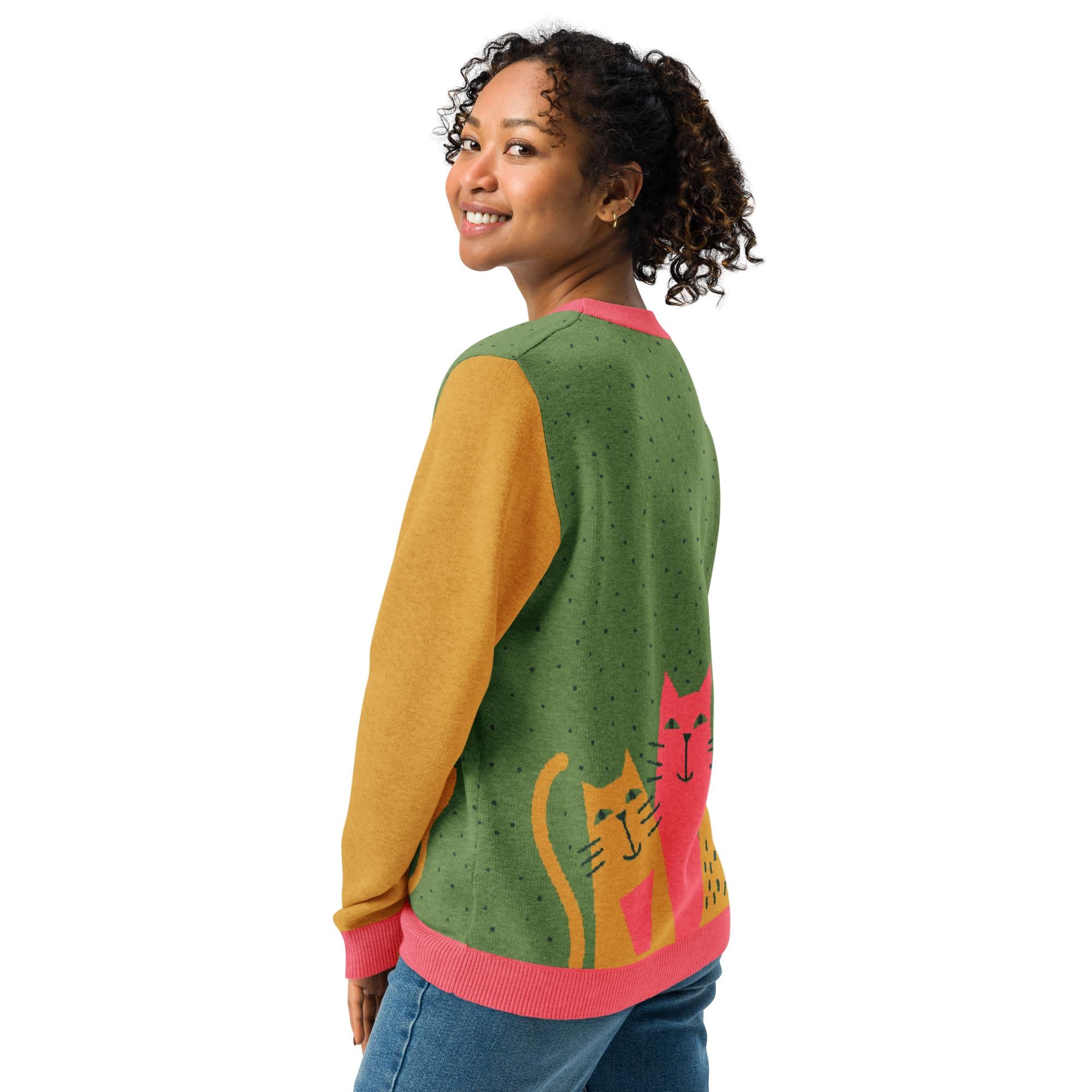 A woman with curly hair is smiling and posing in a colorful sweater with cat designs.