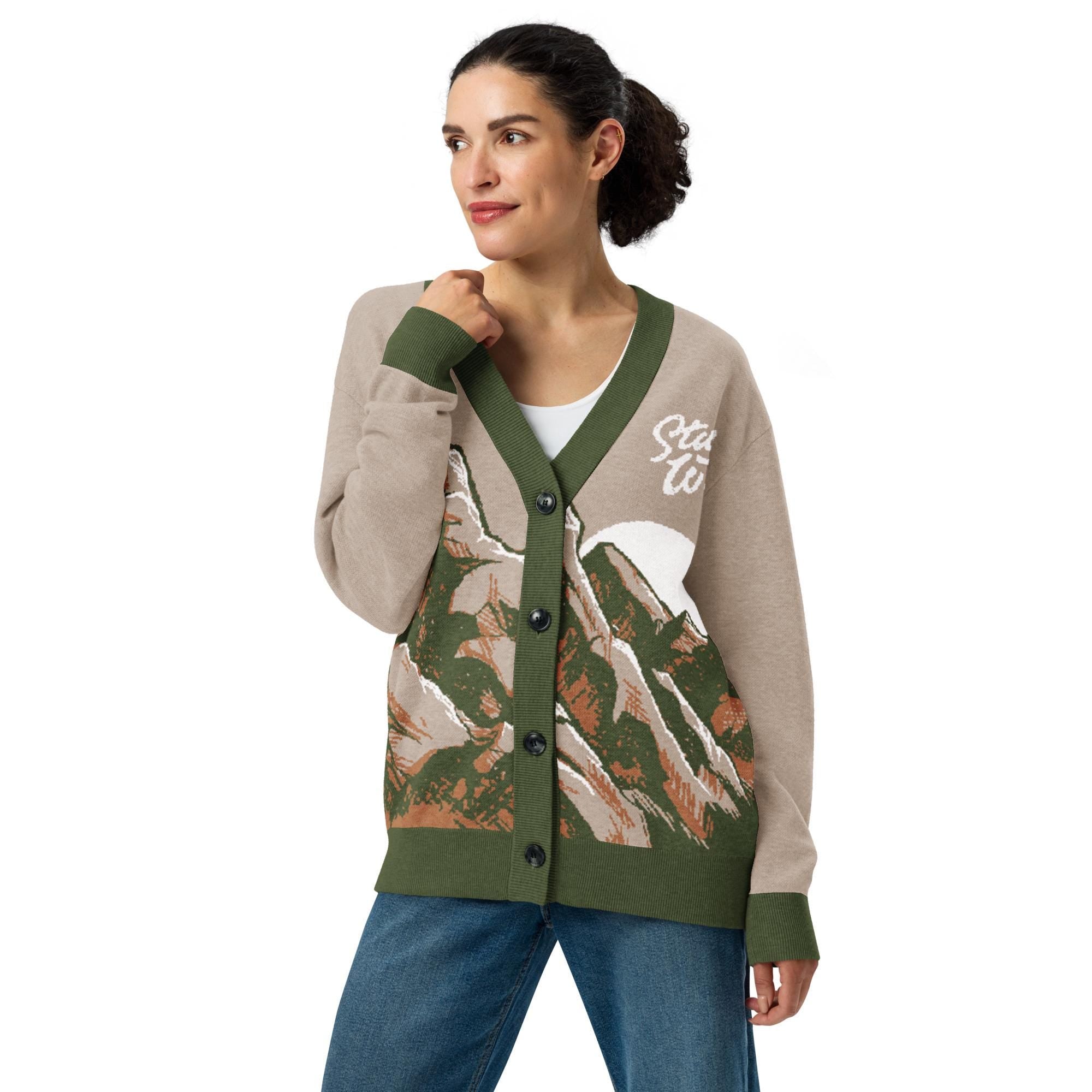 A woman wearing a green and beige cardigan with a mountain landscape design, posing with her hand on her chin.