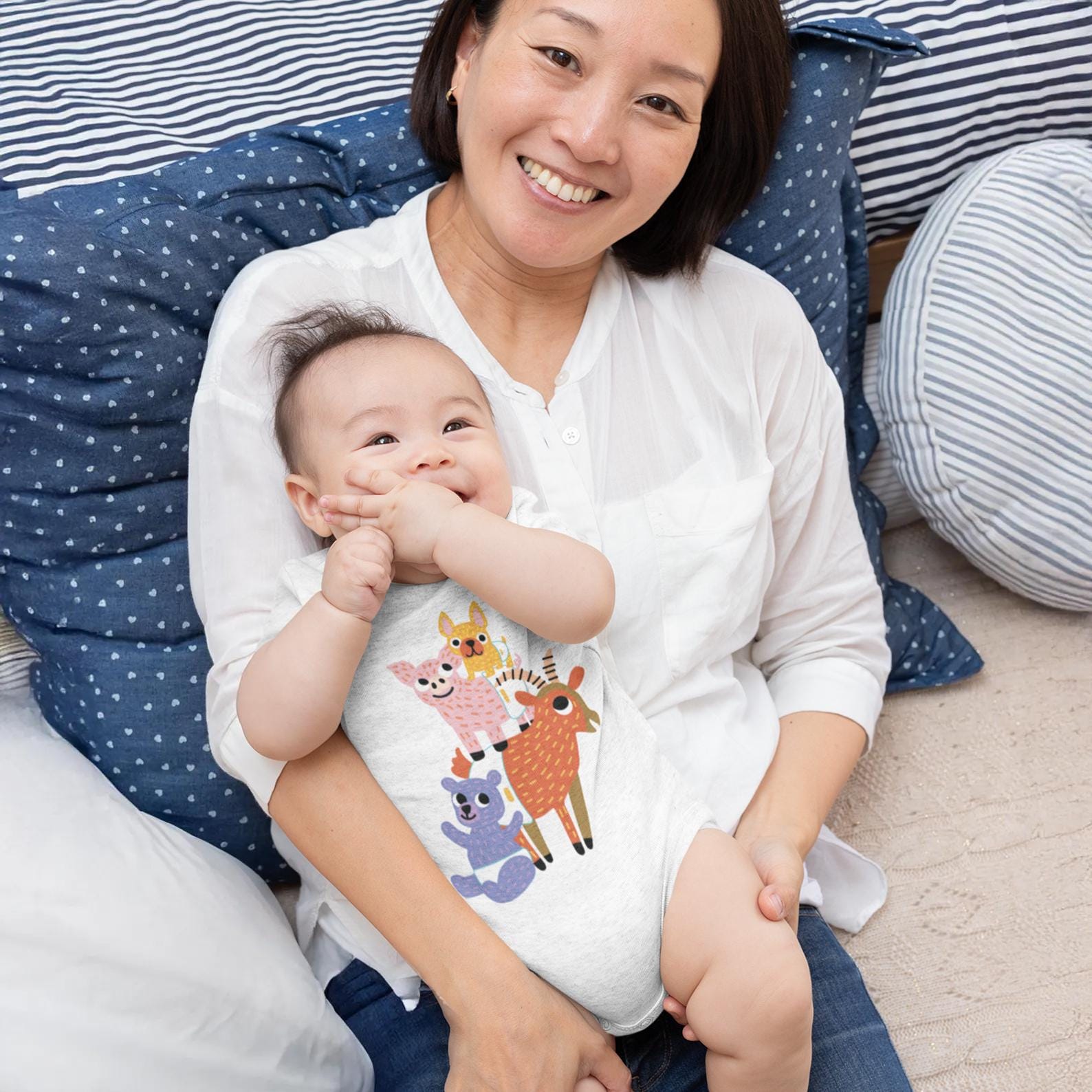 A smiling woman holding a baby in her arms, both sitting on a bed with blue and white pillows.