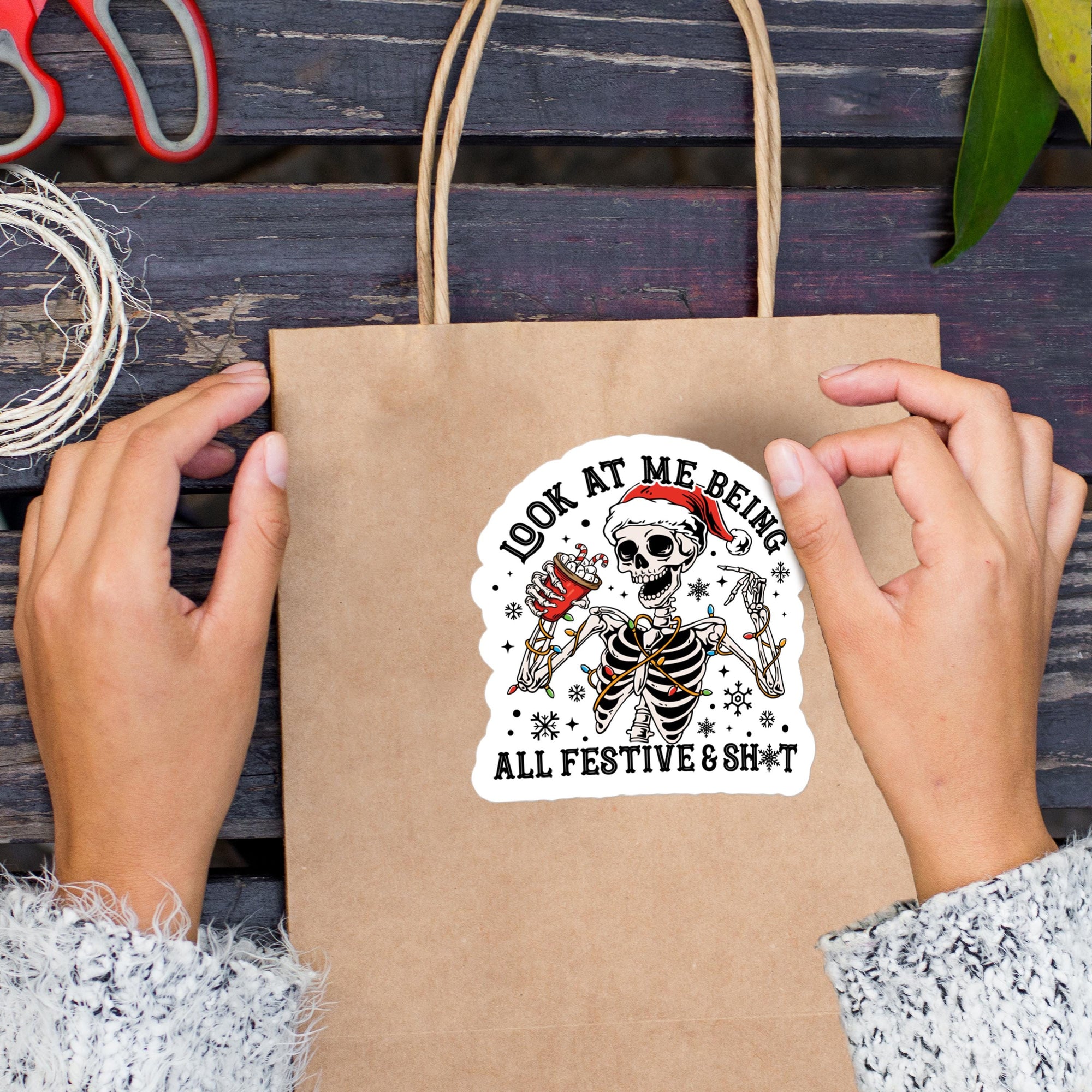 A person&#39;s hands holding a brown paper bag with a sticker featuring a skeleton wearing a Santa hat and the text &quot;Look at me being all festive & short&quot;.