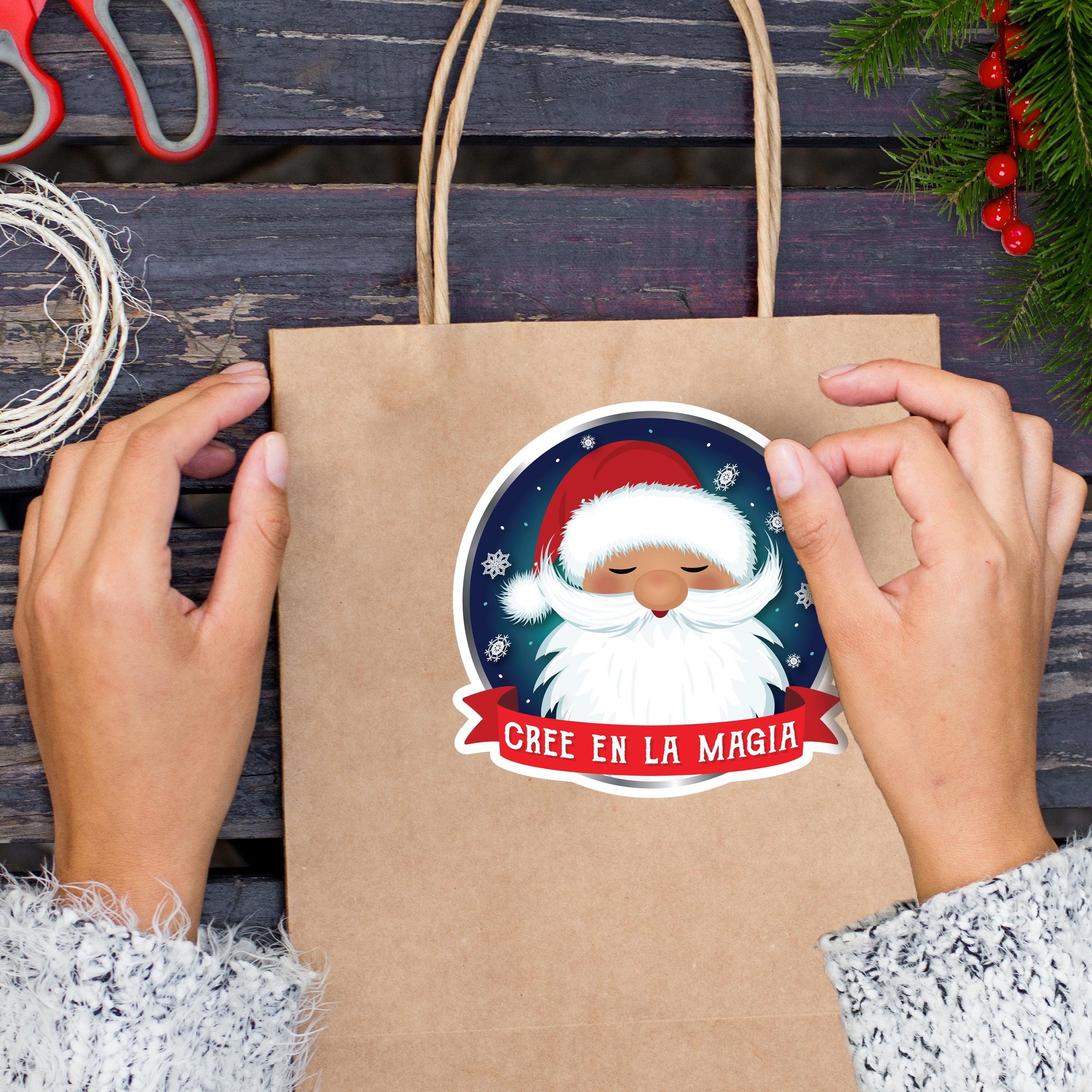 A person&#39;s hands holding a brown paper bag with a Santa Claus sticker on it, surrounded by Christmas decorations and a pair of scissors.