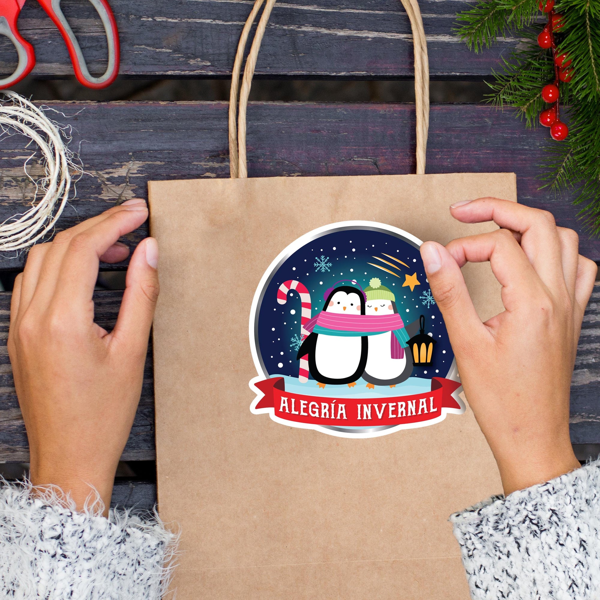 A person&#39;s hands holding a brown paper bag with a sticker featuring a penguin and snowflakes, along with the text &quot;ALEGRÍA INvernal&quot; (Spanish for &quot;Christmas invernal&quot; or &quot;Christmas invernal&quot;).